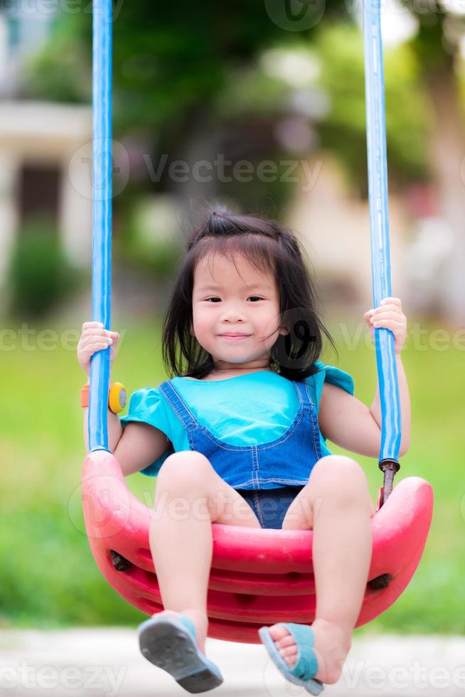 chica asiática juega un columpio rojo en el patio de recreo. en verano o primavera. niños lindos sonriendo dulce y brillante. el fondo es césped verde borroso. niño de 3 años. concepto de actividades de ocio y relajación. foto