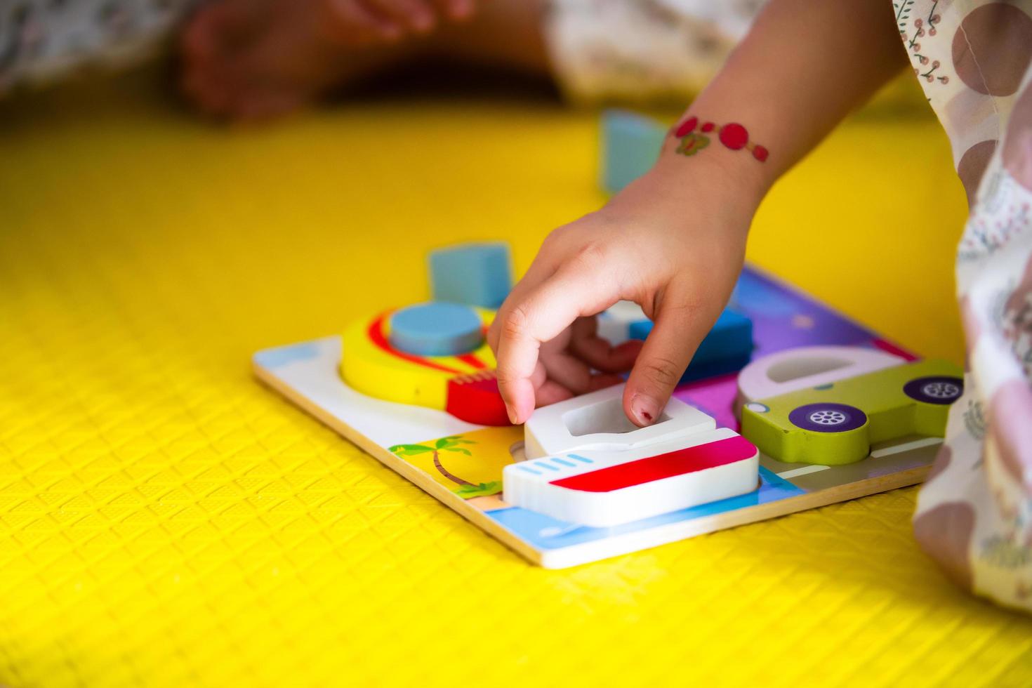 The little girl's hands are playing wooden puzzles to match the shapes to their correct boxes. On a cloudy day. Floor yellow. On the wrist of the little child, put on tattoos. photo