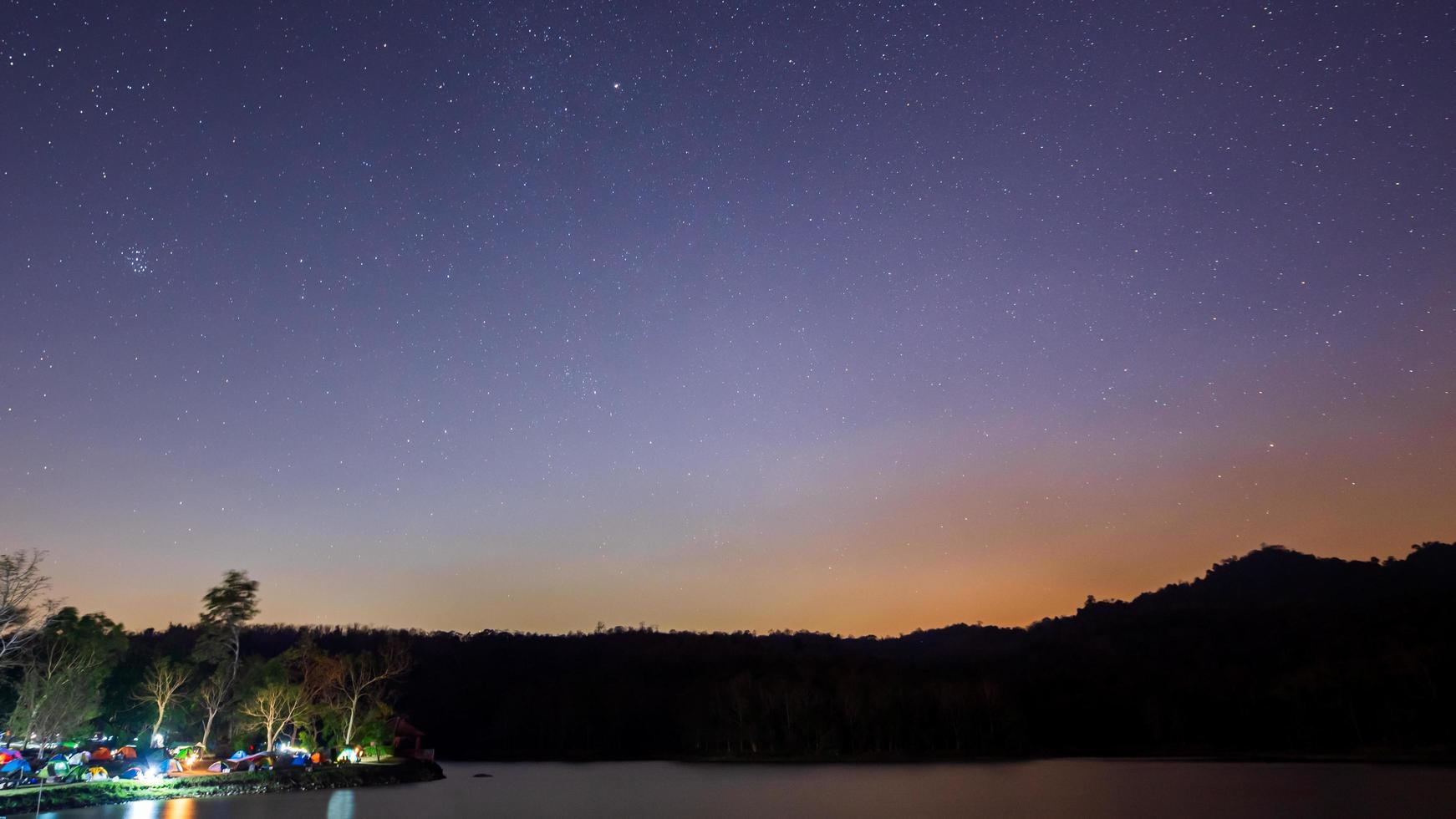 Shadow mountain range, orange-yellow rim light, along the night ridge, stars in the opposite direction of the Milky Way. Tourists camped in tents under the trees at the lighted corner photo