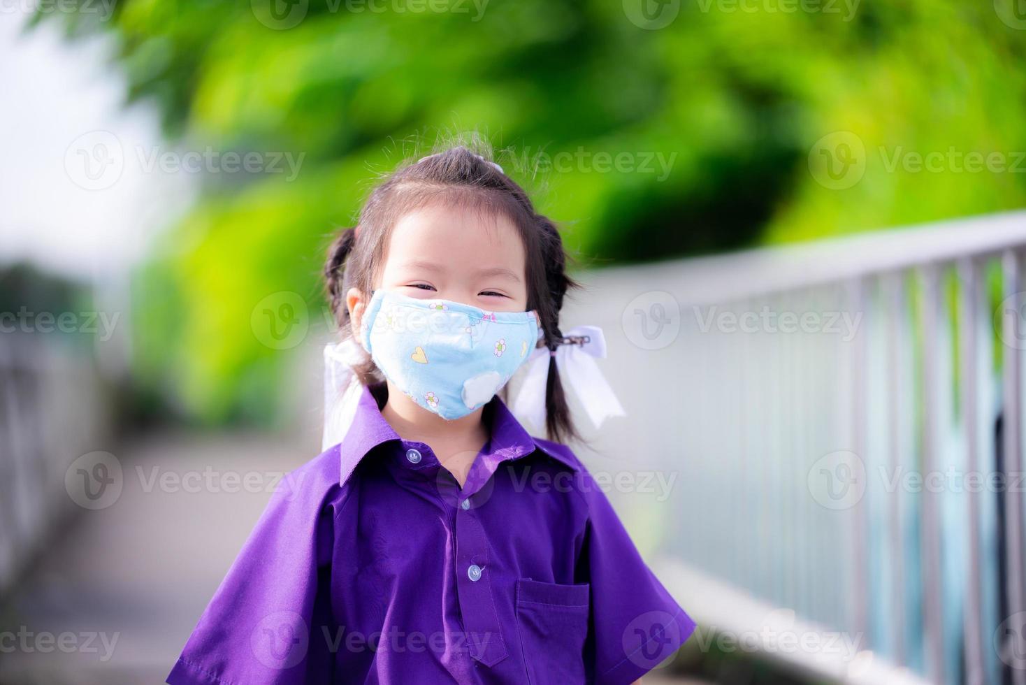 niño feliz usa máscara de tela cuando va a la escuela. chica asiática sonríe dulce debajo de la mascarilla para prevenir la infección por virus y evitar el polvo y el humo cuando está en público. niño de 3 años de edad de nueva vida normal foto