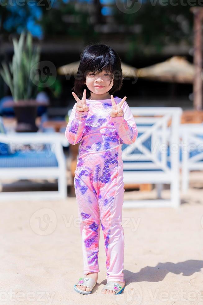 vertical imaged. Happy Asian cute girl standing and posting take a picture. Child looking at camera. Sweet smiling. At the beach. Summer time. Children wearing swimsuit. Baby aged 4-5 years old. photo