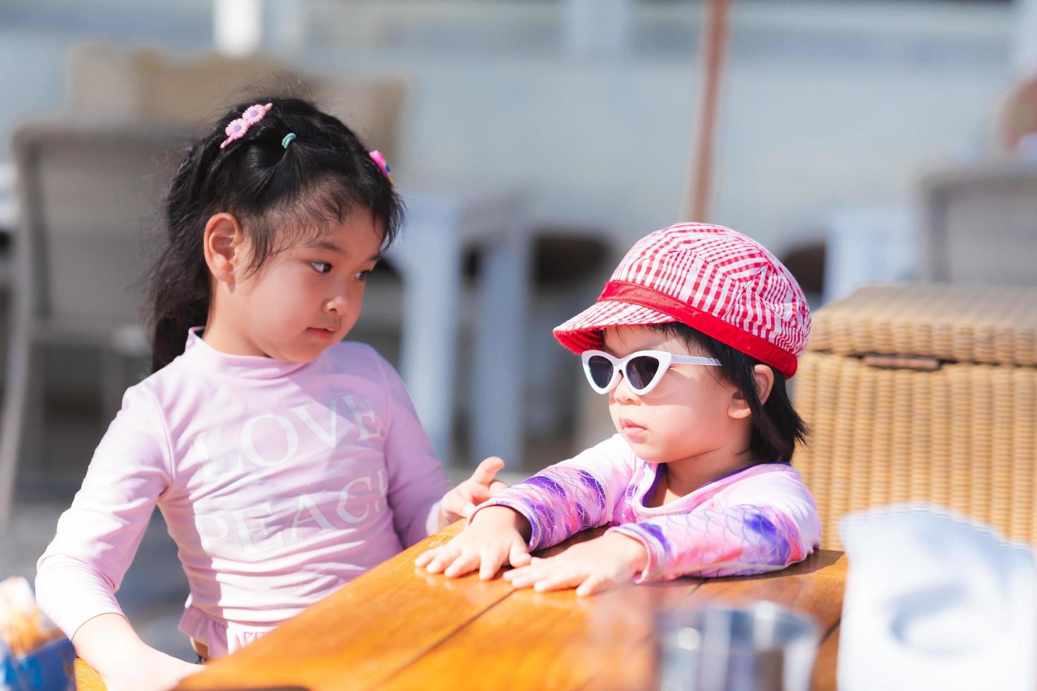 adorables dos chicas asiáticas se ponen de pie y hablan. el niño usa gafas de sol y un sombrero. los niños van al mar. mujeres usan trajes de baño. niño se pega a una mesa en un restaurante. bebé de 4-5 años. foto