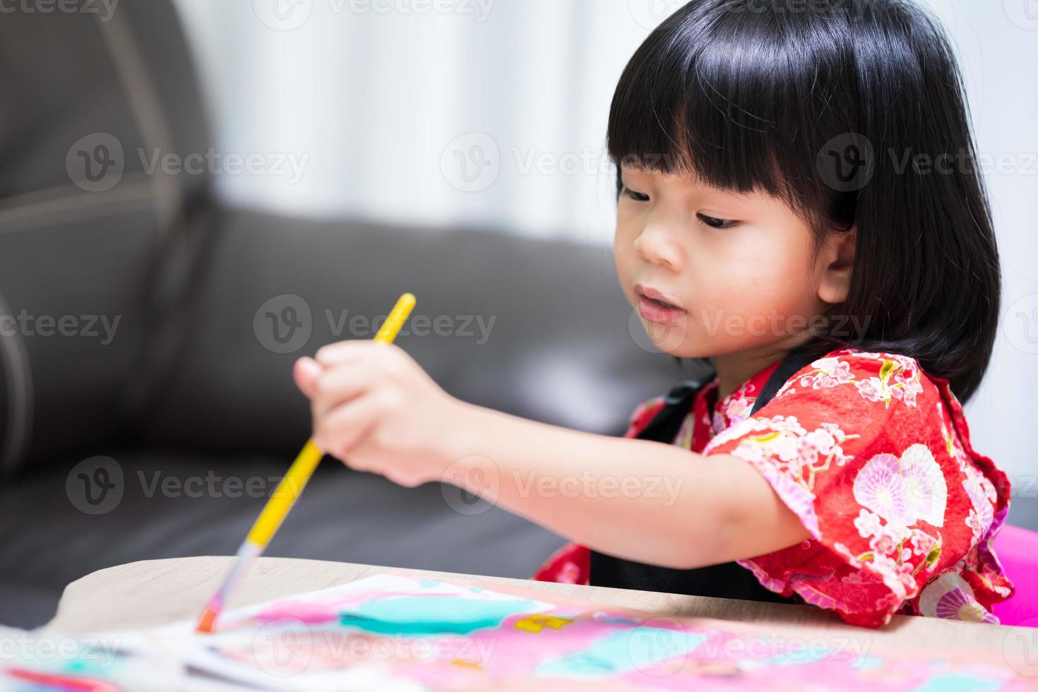 Child holding brush in her left hand is painting water color on paper for the artwork placed on table. Children have fun learning and building imaginations. Happy kid wear black apron uniforms. photo