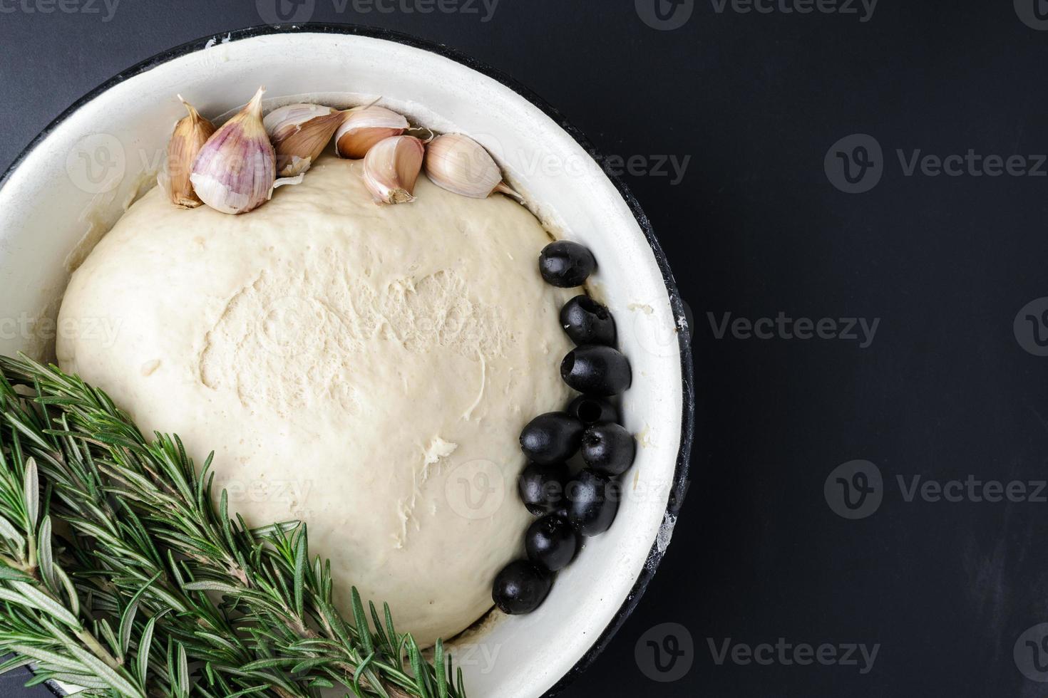 Raw dough in a white bowl, sprigs of rosemary, olives and garlic on a dark background. photo