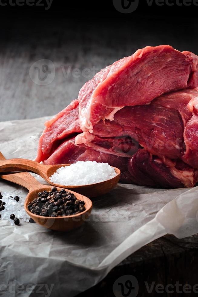 un gran trozo de carne de cerdo, sal y pimienta en cucharas de madera sobre una mesa de madera sobre un fondo oscuro. foto