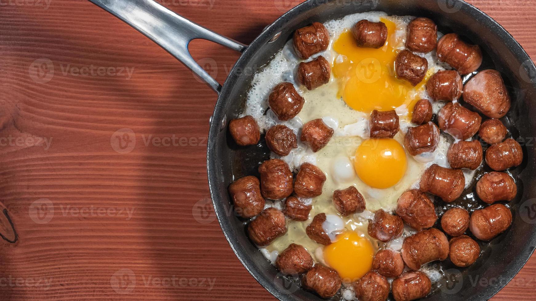 Banner with fried sausages with chicken eggs in process of frying in a frying pan at the red wood table with copy space for text. photo