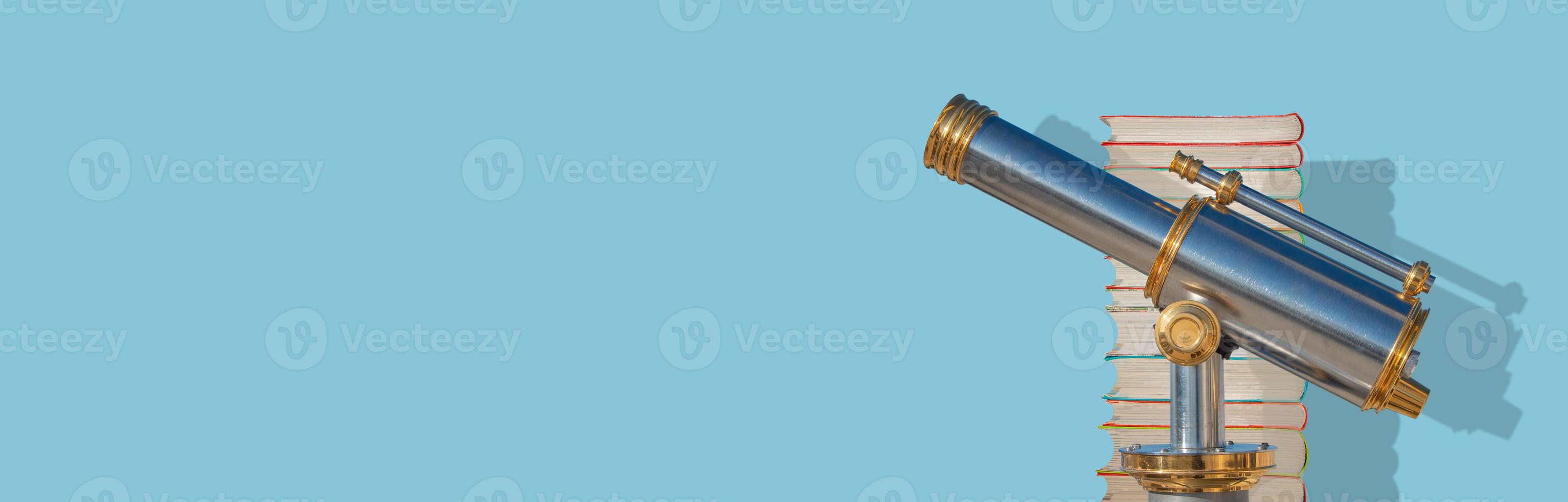 Banner with an old antique metal telescope standing near a stack of books at solid blue background with copy space and shadow, details, closeup. Concept exploration, education and travel. photo