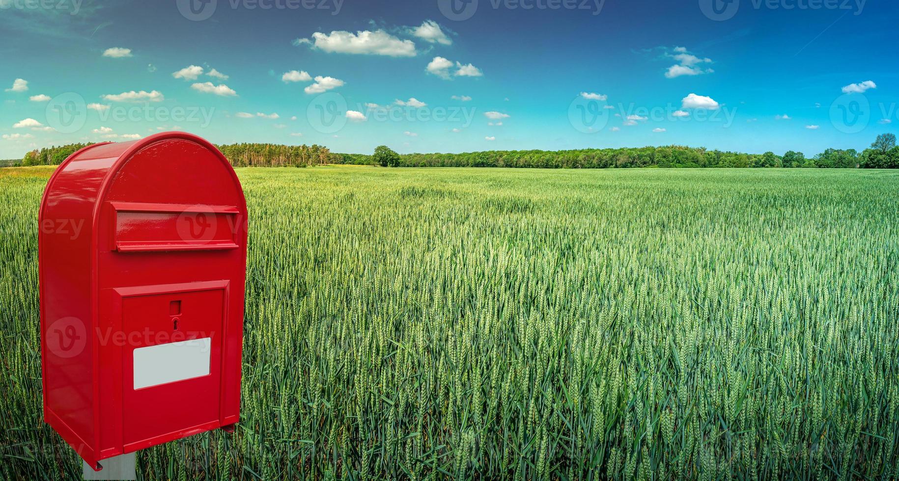 Big red modern postbox with white empty note space for address is standing outdoor in front of beautiful countryside landscape background with farm green wheat field and blue sky. photo