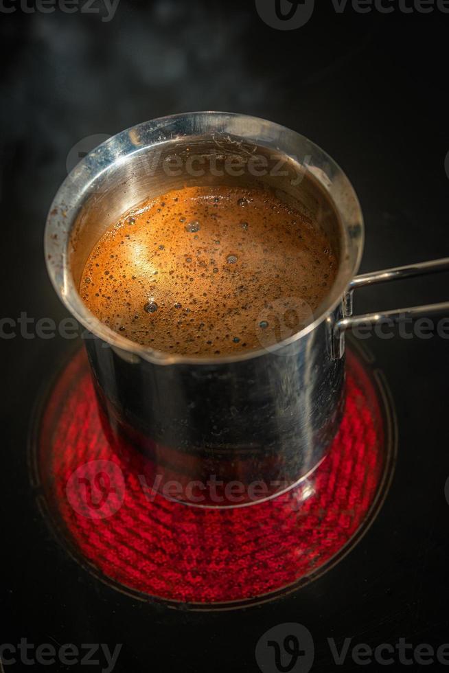 Stainless steel cezve with coffee in process of making standing at hot and red electrical stove in darkness, closeup, details photo