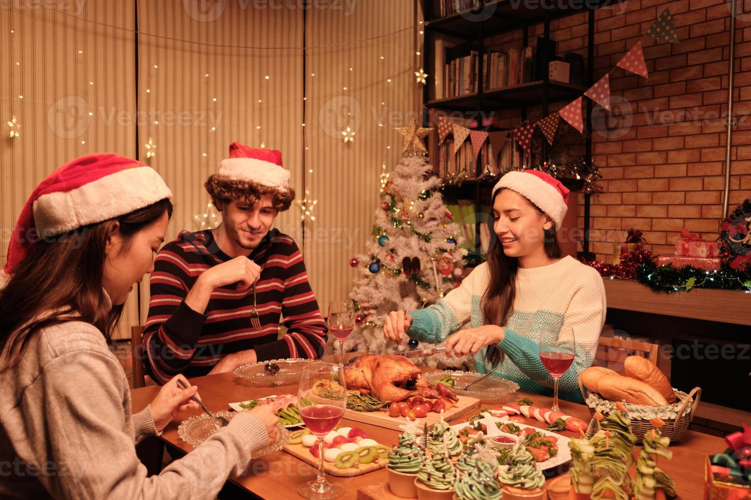 Friends enjoy fun eating diner at table with specials foods, young woman cutting roasted turkey at home's dining room, decorated with ornaments, Christmas festival, and New Year celebration party. photo