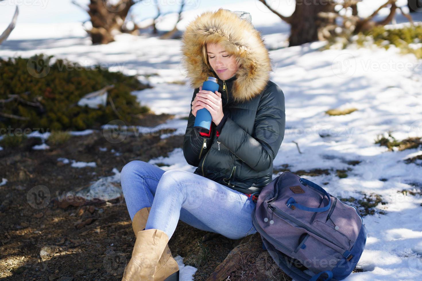 mujer bebiendo algo caliente de una botella termo de metal sentada sobre una roca en las montañas nevadas. foto