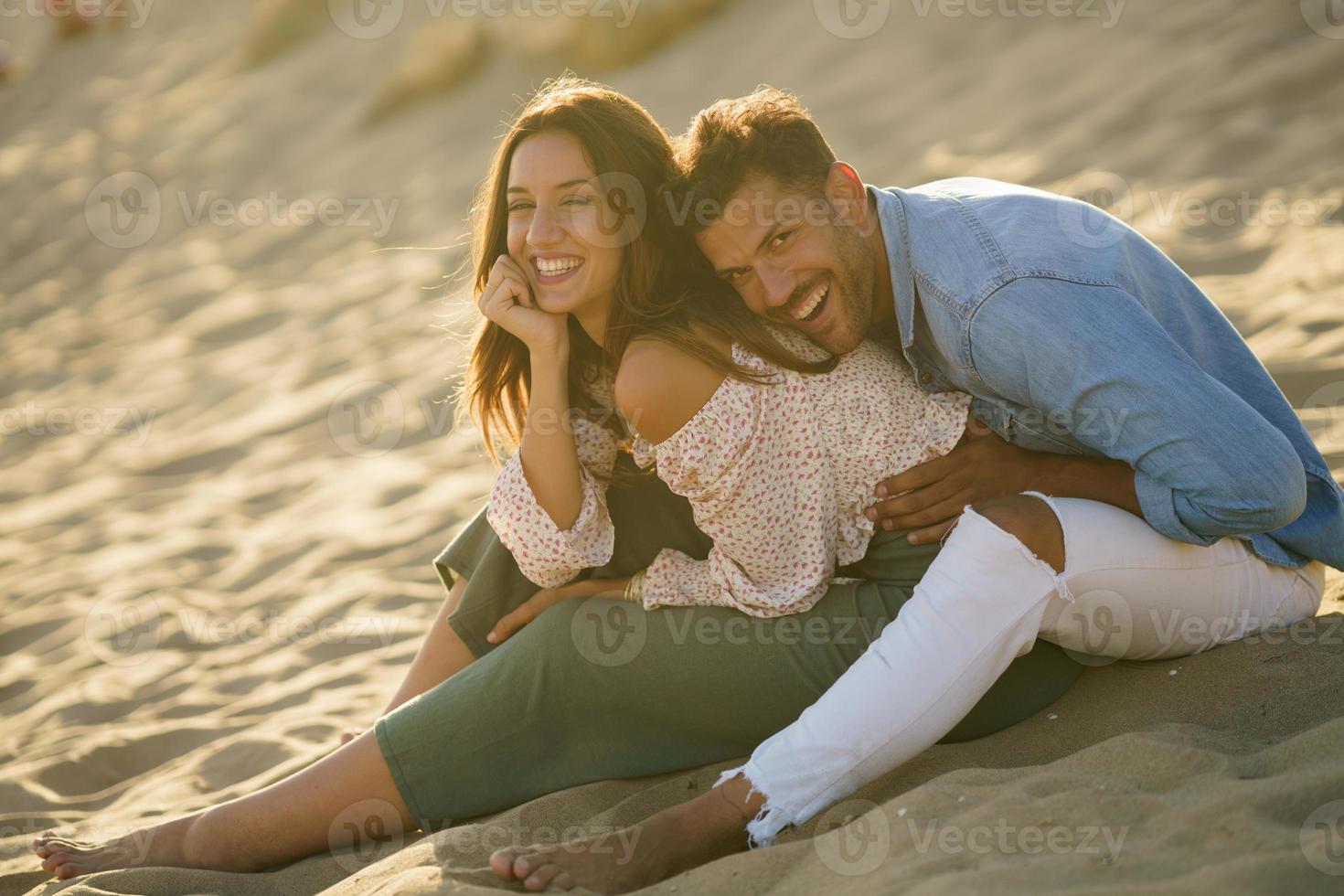 Pareja joven riendo juntos sentados en la arena de la playa foto