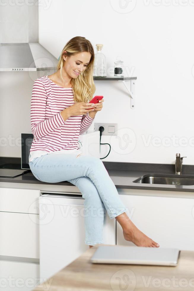 Happy beautiful woman using her smartphone sitting in the kitchen at home photo