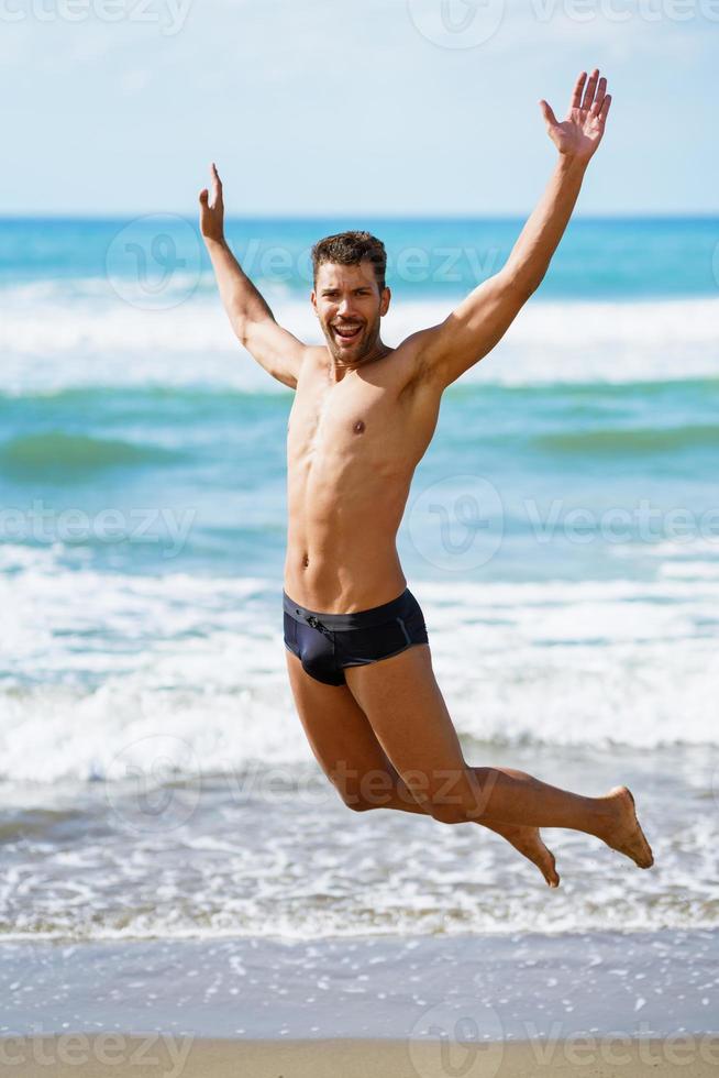 Young man with beautiful body in swimwear jumping on a tropical beach. photo
