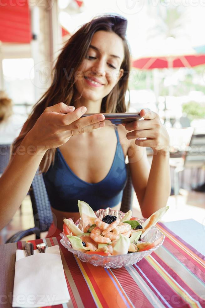 Mujer joven fotografiando su ensalada con un teléfono inteligente mientras está sentado en un restaurante foto