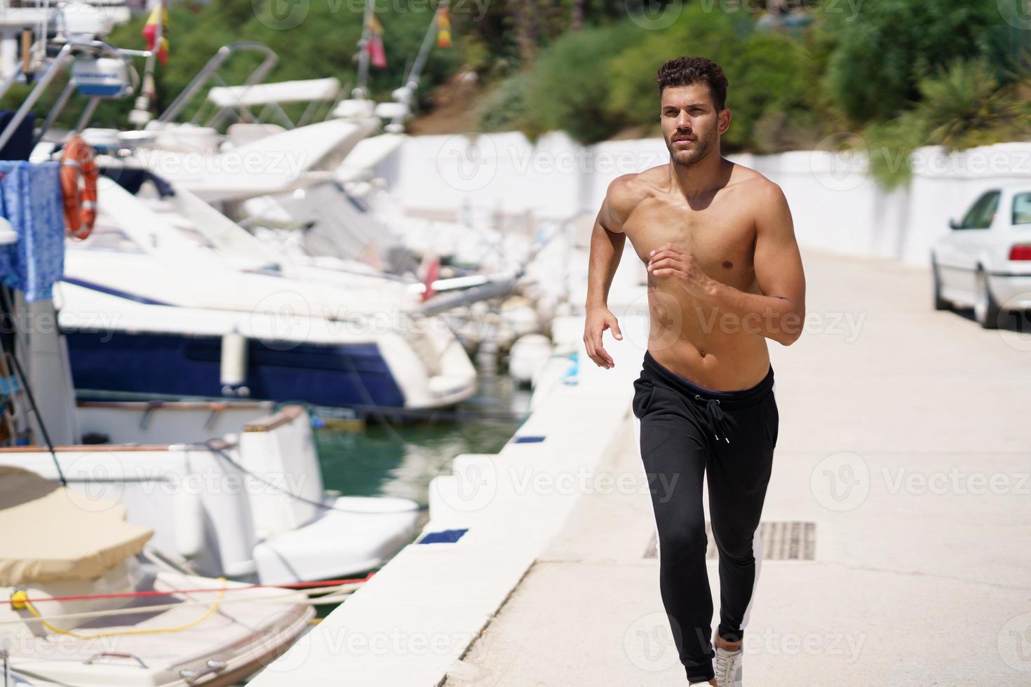 Young man with an athletic body running shirtless through a harbour. photo