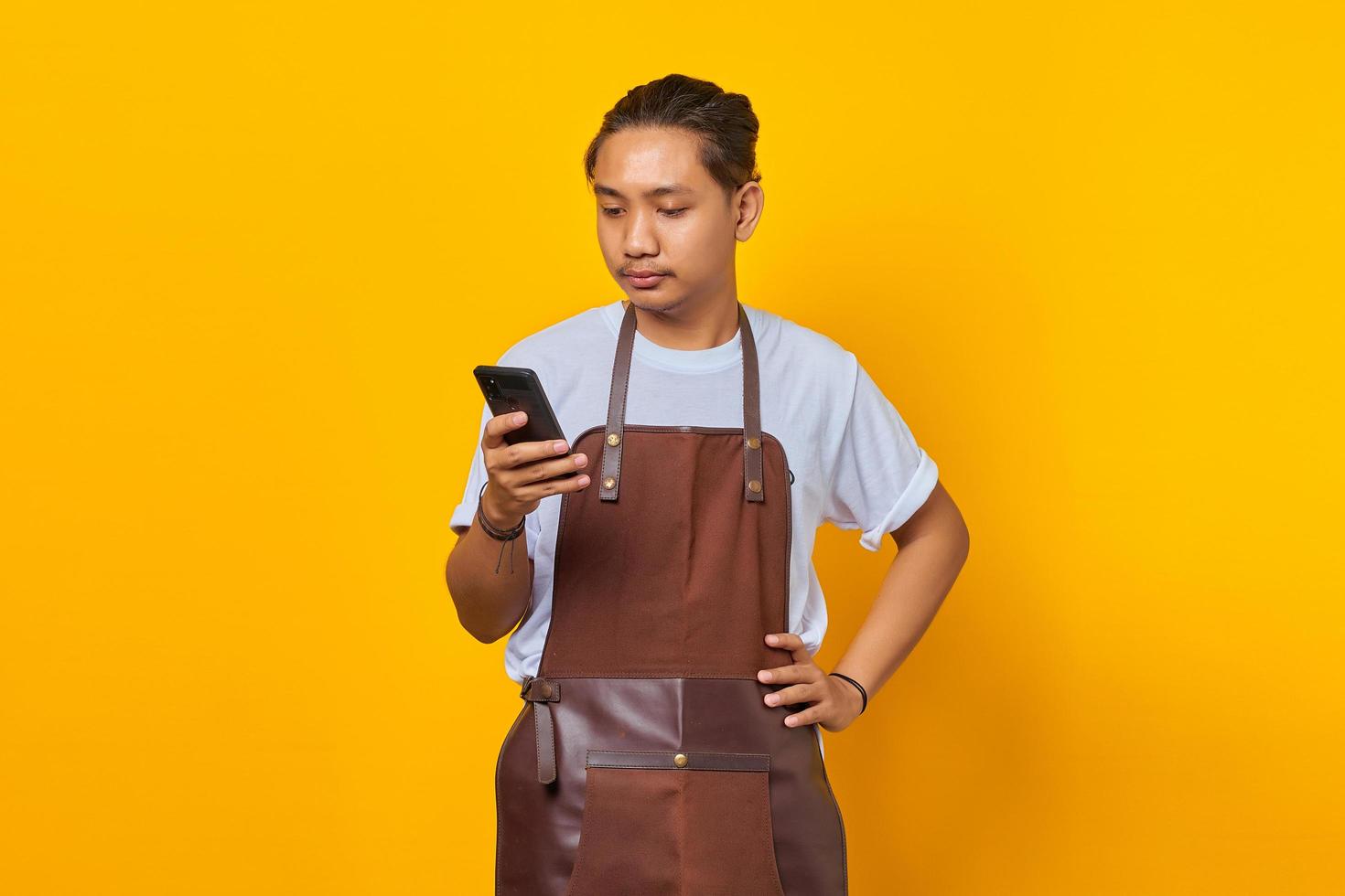 Portrait of handsome asian man wearing apron looking unhappy reading incoming messages on mobile on yellow background photo