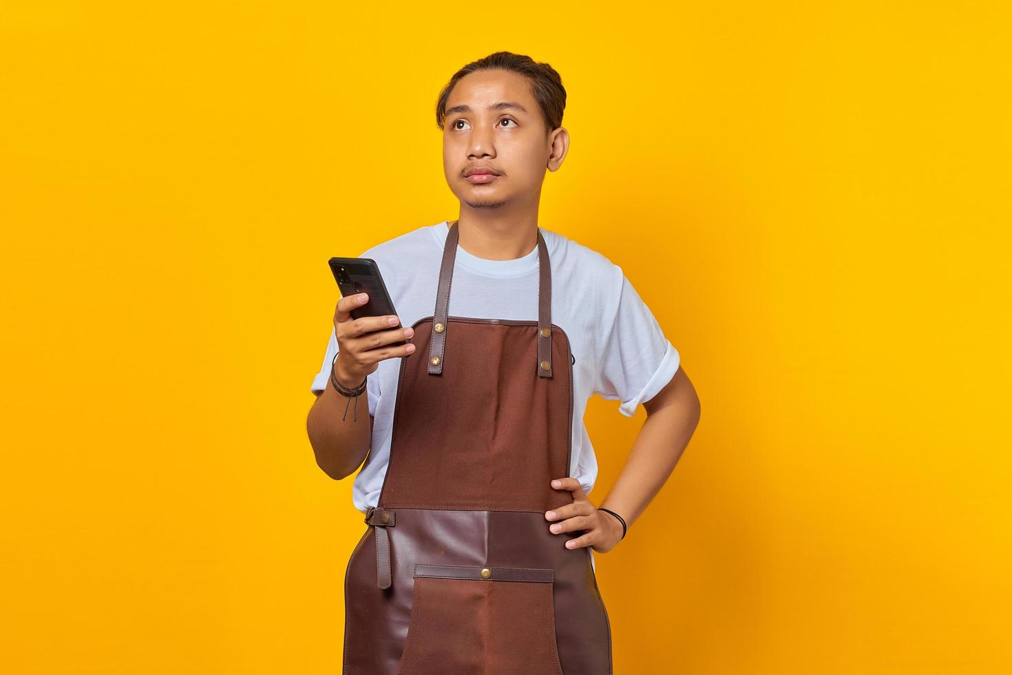 Surprised handsome barista holding cell phone and looking at empty space on yellow background photo