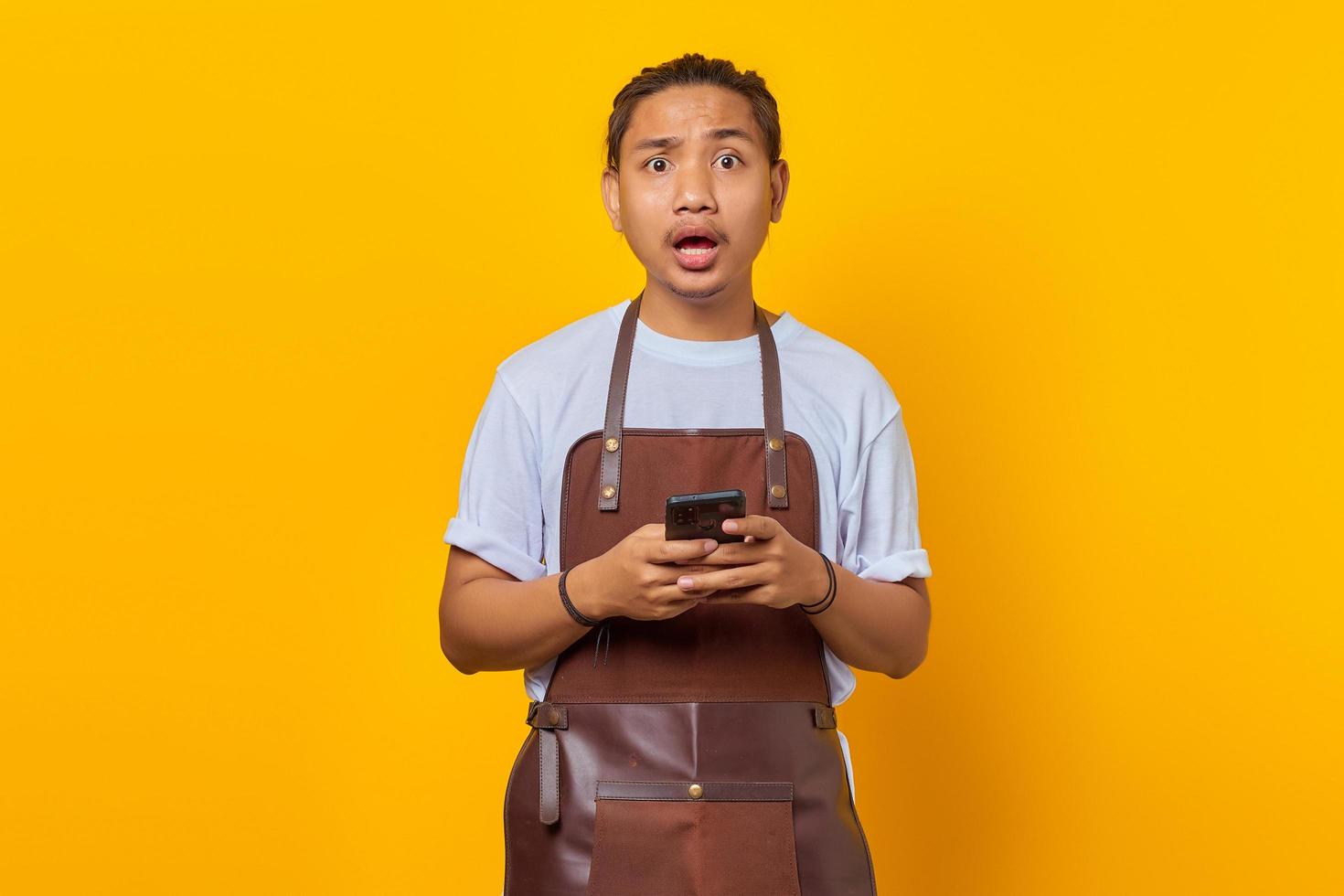 Portrait of Asian young man wearing apron looking surprised and holding smartphone on yellow background photo