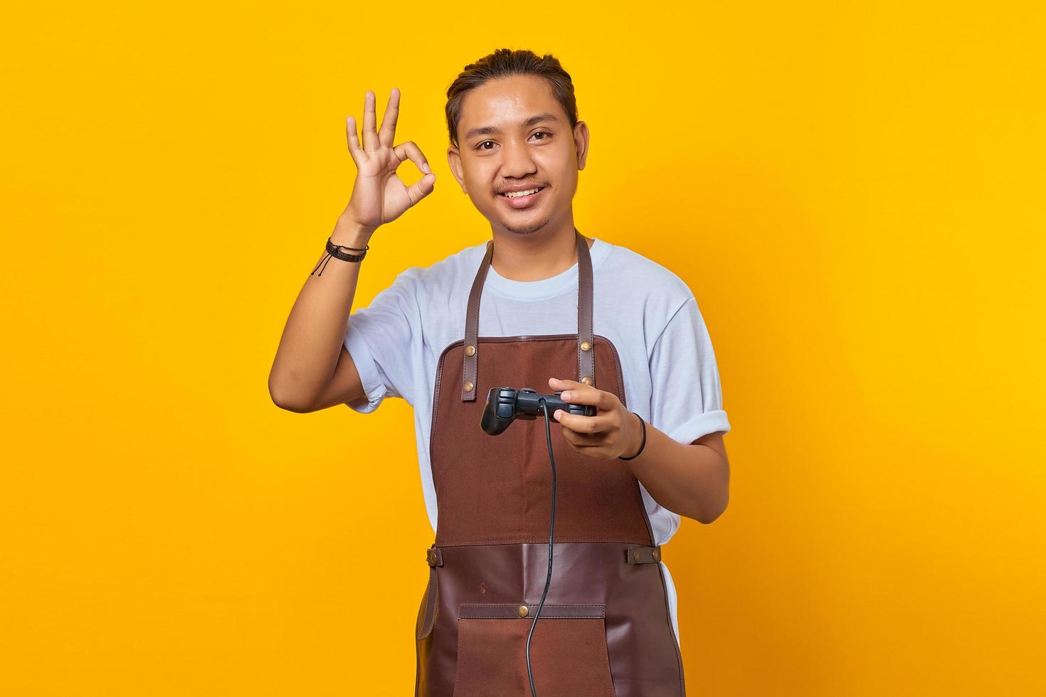 Portrait of cheerful handsome Asian young man wearing apron holding game controller and confident showing ok gesture over yellow background photo