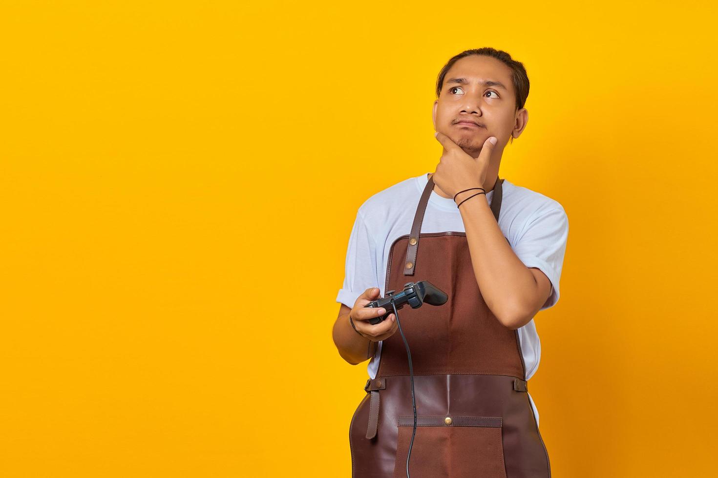 Young man wearing apron holding game controller looking aside with doubtful and skeptical expression on yellow background photo