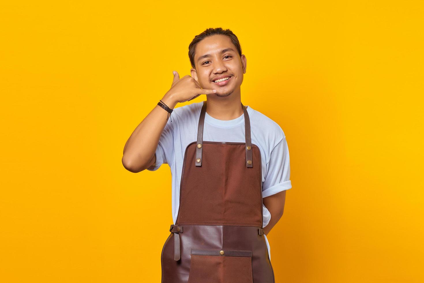Portrait of smiling Asian young man wearing apron making call sign on yellow background photo