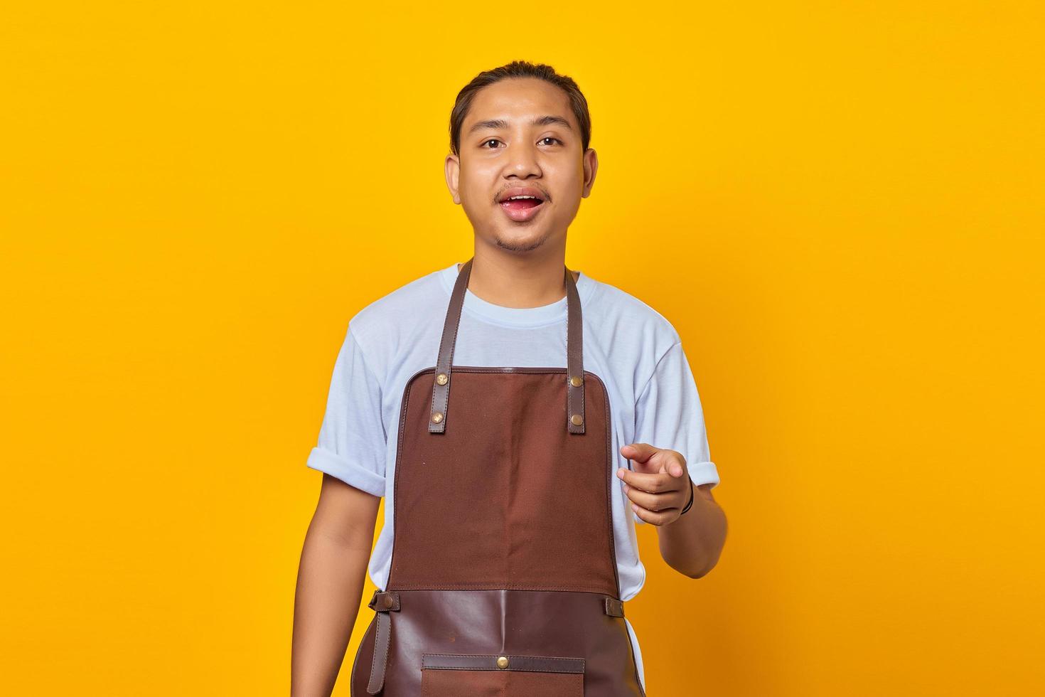 Retrato de sonriente joven asiático vistiendo delantal apuntando a la cámara con el dedo sobre fondo amarillo foto