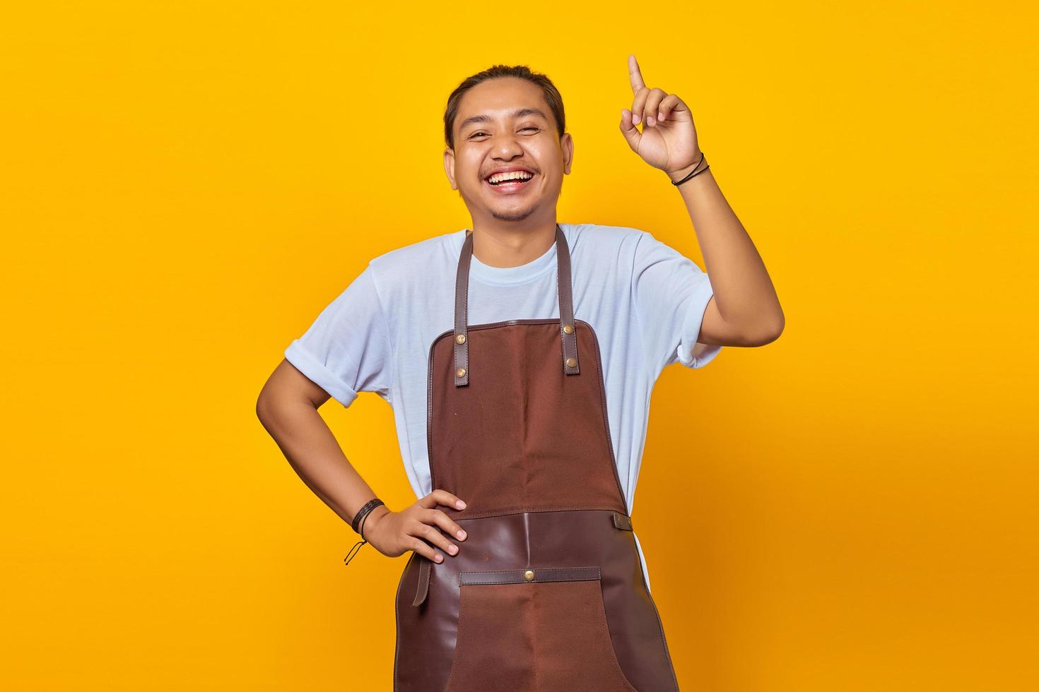 Cheerful handsome Asian young man wearing apron pointing up to copy space isolated on yellow background photo