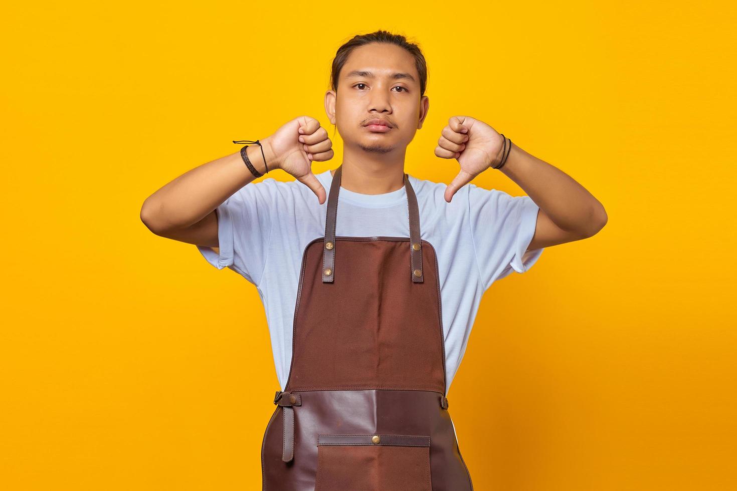 Portrait of attractive Asian young man wearing apron unhappy and angry showing rejection with thumbs down gesture photo