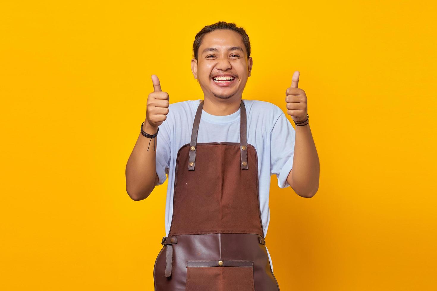 Alegre joven barista guapo mostrando los pulgares hacia arriba o signo de aprobación aislado sobre fondo amarillo foto