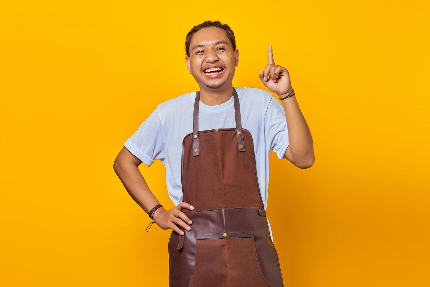 Cheerful handsome Asian young man wearing apron pointing up to copy space isolated on yellow background photo