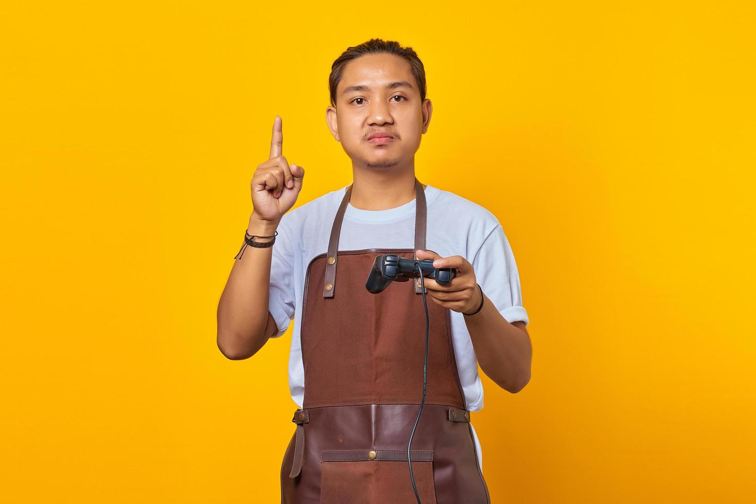 Retrato de un apuesto joven asiático vistiendo delantal sosteniendo un controlador de juego con una gran idea aislada sobre fondo amarillo foto