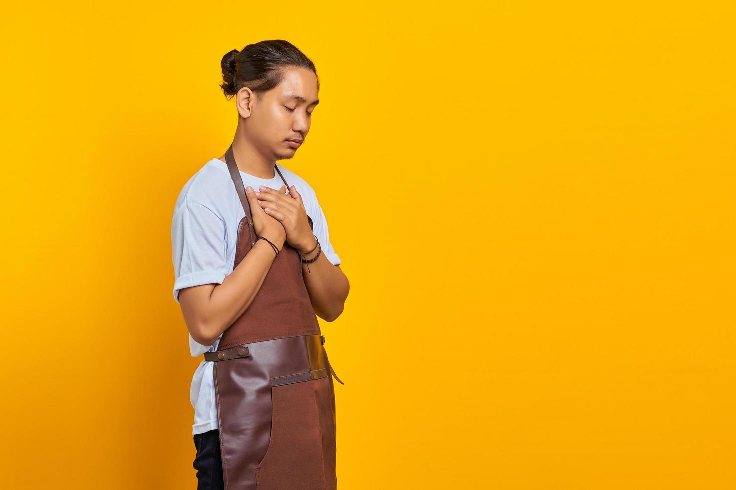 Portrait of handsome Asian young man wearing apron smiling with hands on chest with closed eyes and grateful face on yellow background photo
