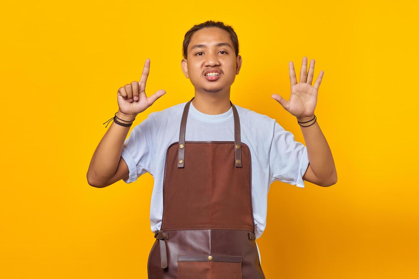 Portrait of confident handsome Asian young man wearing apron pointing with finger number seven with angry expression photo