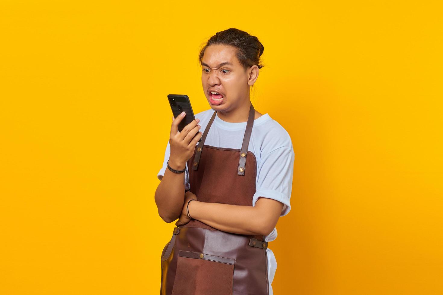 Retrato de joven asiático vistiendo delantal con enojo mientras habla por teléfono inteligente sobre fondo amarillo foto