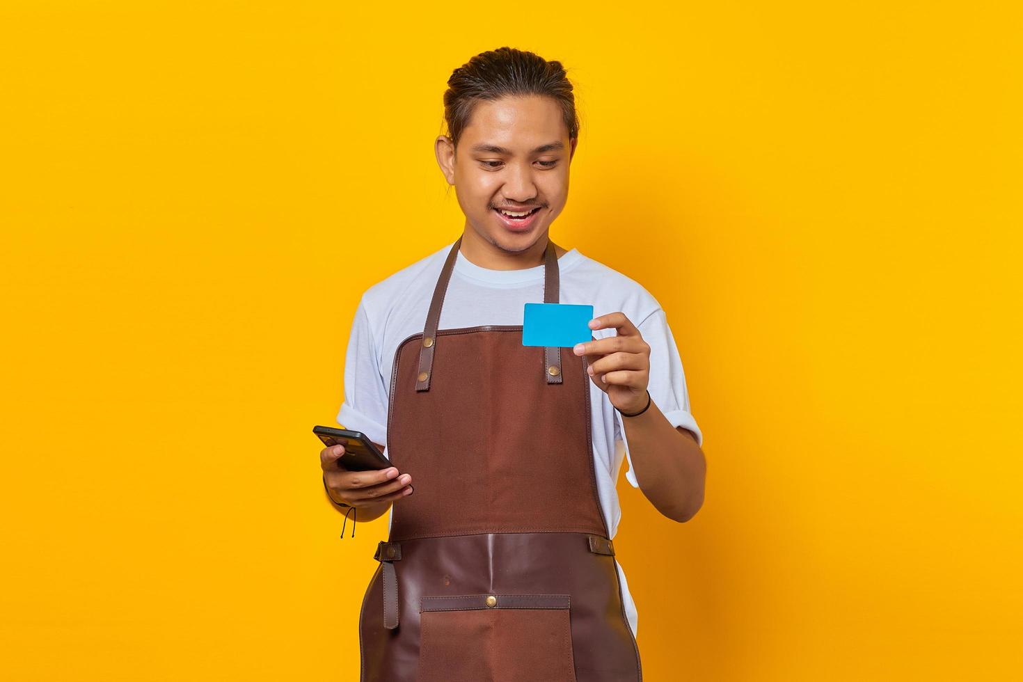 Portrait of cheerful handsome barista looking at credit card and holding smartphone isolated on yellow background photo