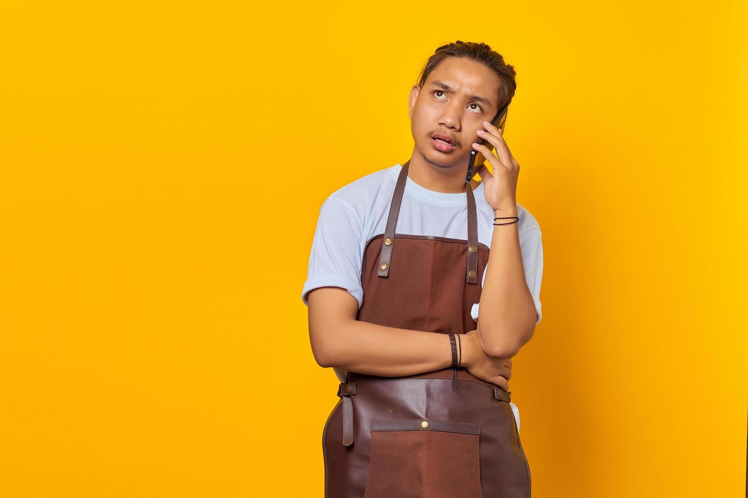 Portrait of unhappy young Asian man while receiving incoming call on smartphone on yellow background photo