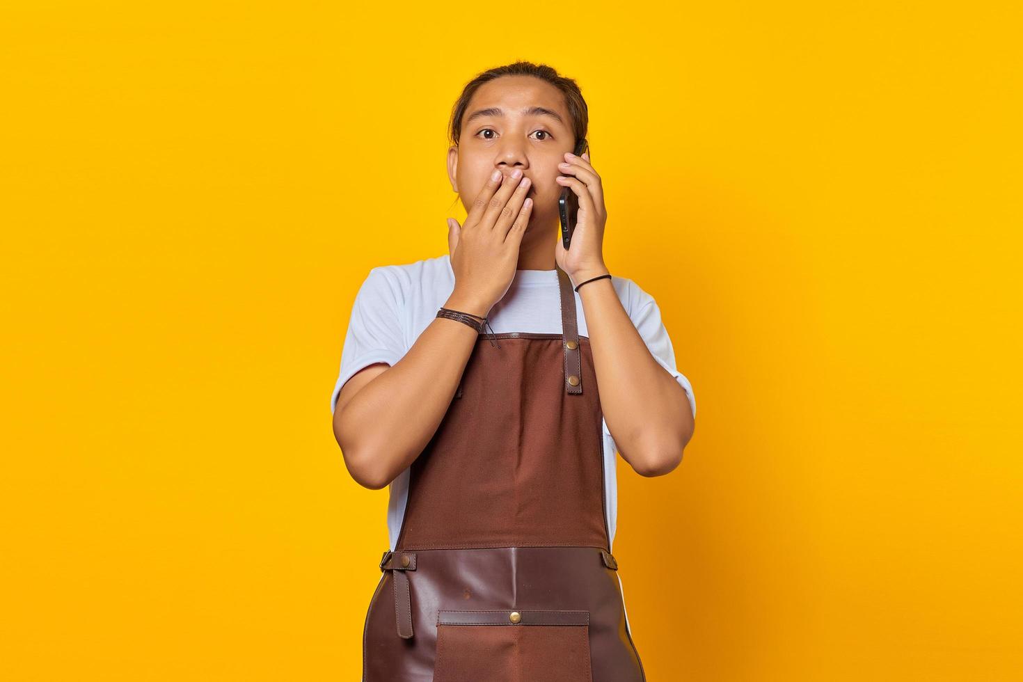 Shocked asian man covering mouth with hands and holding smartphone over yellow background photo