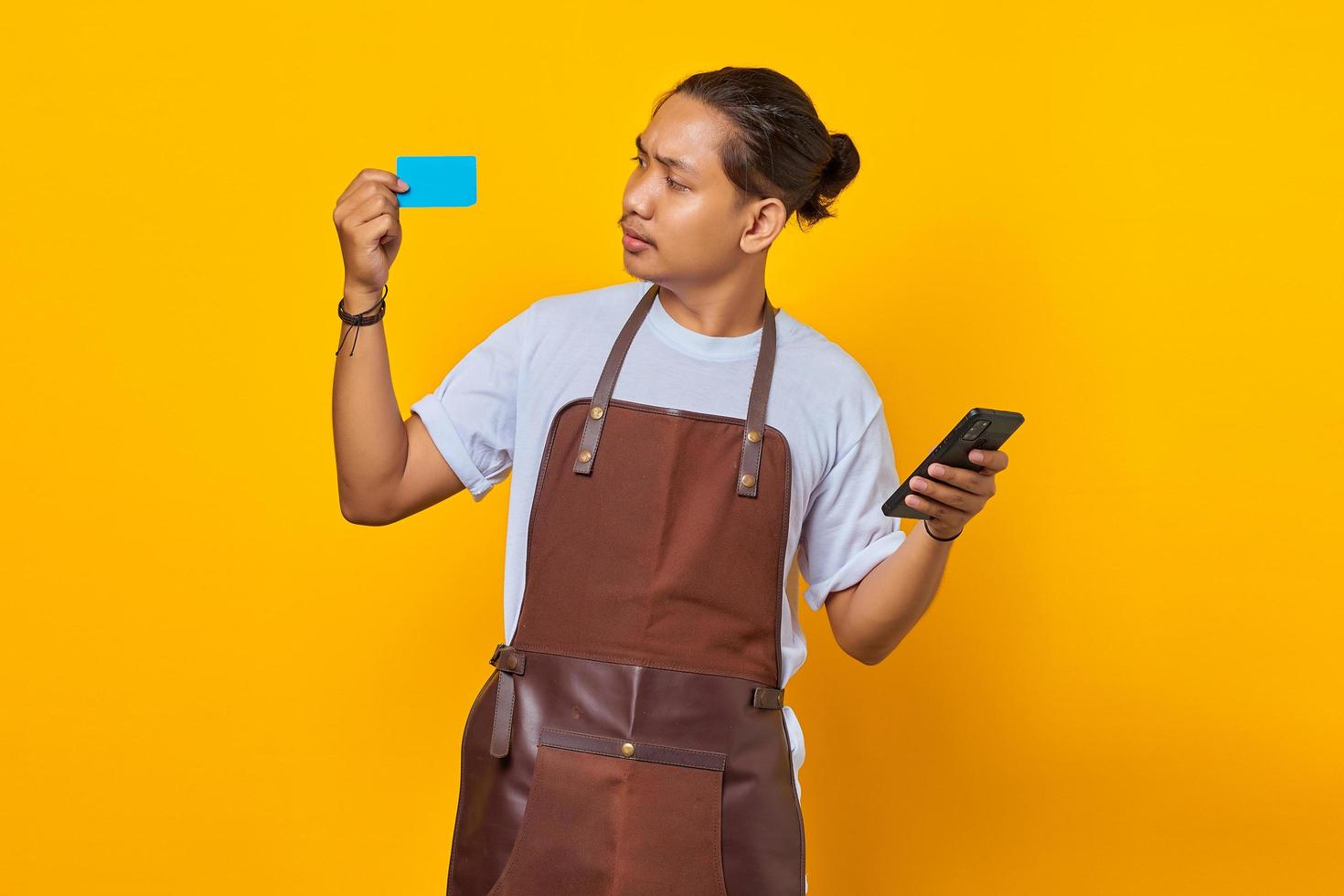 Surprised barista showing credit card to pay and holding smartphone isolated on yellow background photo