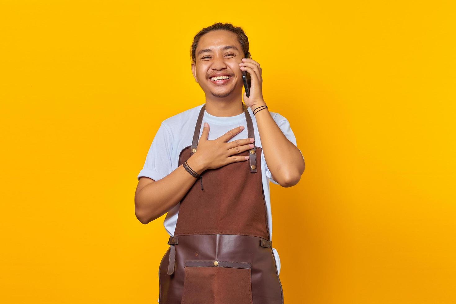 Portrait of cheerful handsome Asian young man wearing apron talking on mobile and hands on chest isolated on yellow background photo
