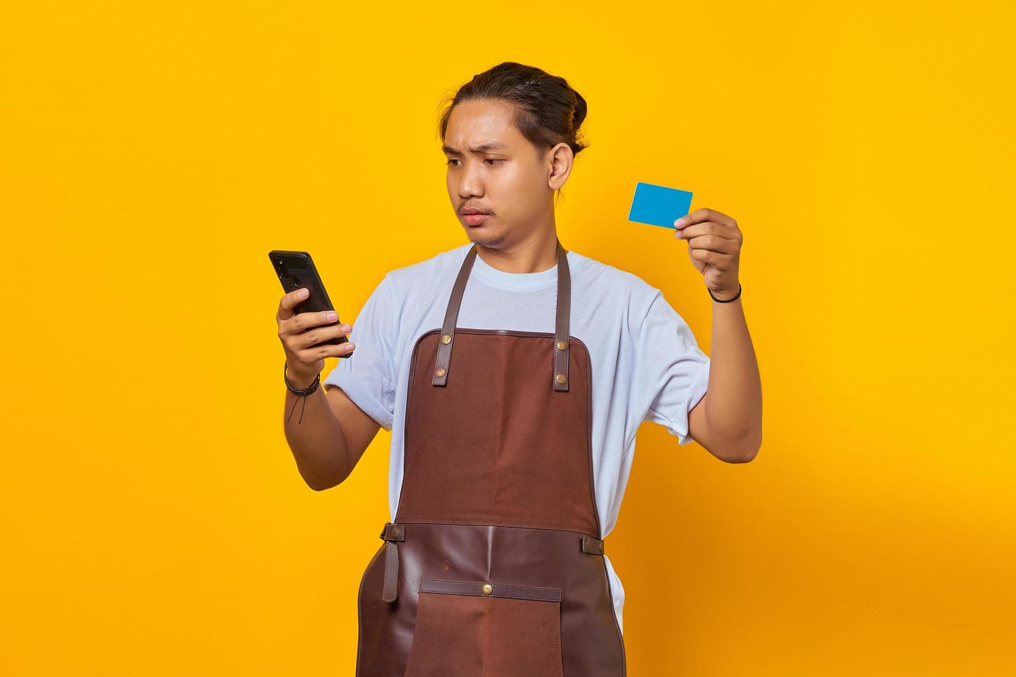 Surprised barista showing credit card to pay and holding smartphone isolated on yellow background photo