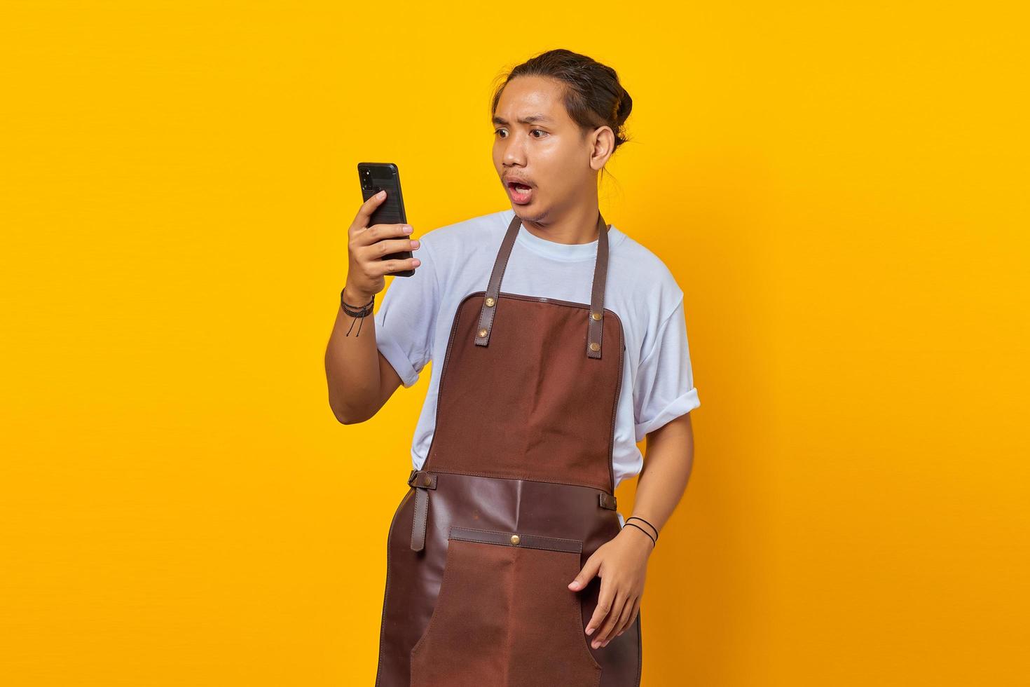 Portrait of Asian young man wearing apron surprised to see incoming message on smartphone on yellow background photo