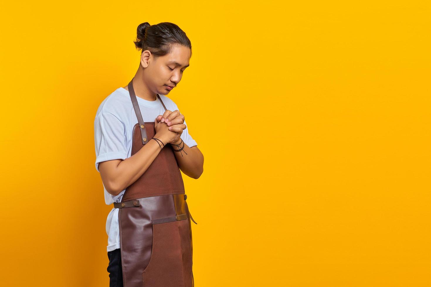 Portrait of handsome Asian young man wearing apron praying, believing in master on yellow background photo