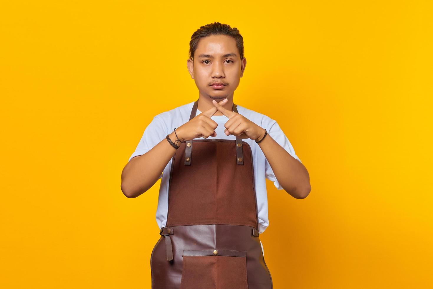 Serious asian young man showing refusal gesture with crossed fingers isolated on yellow background photo