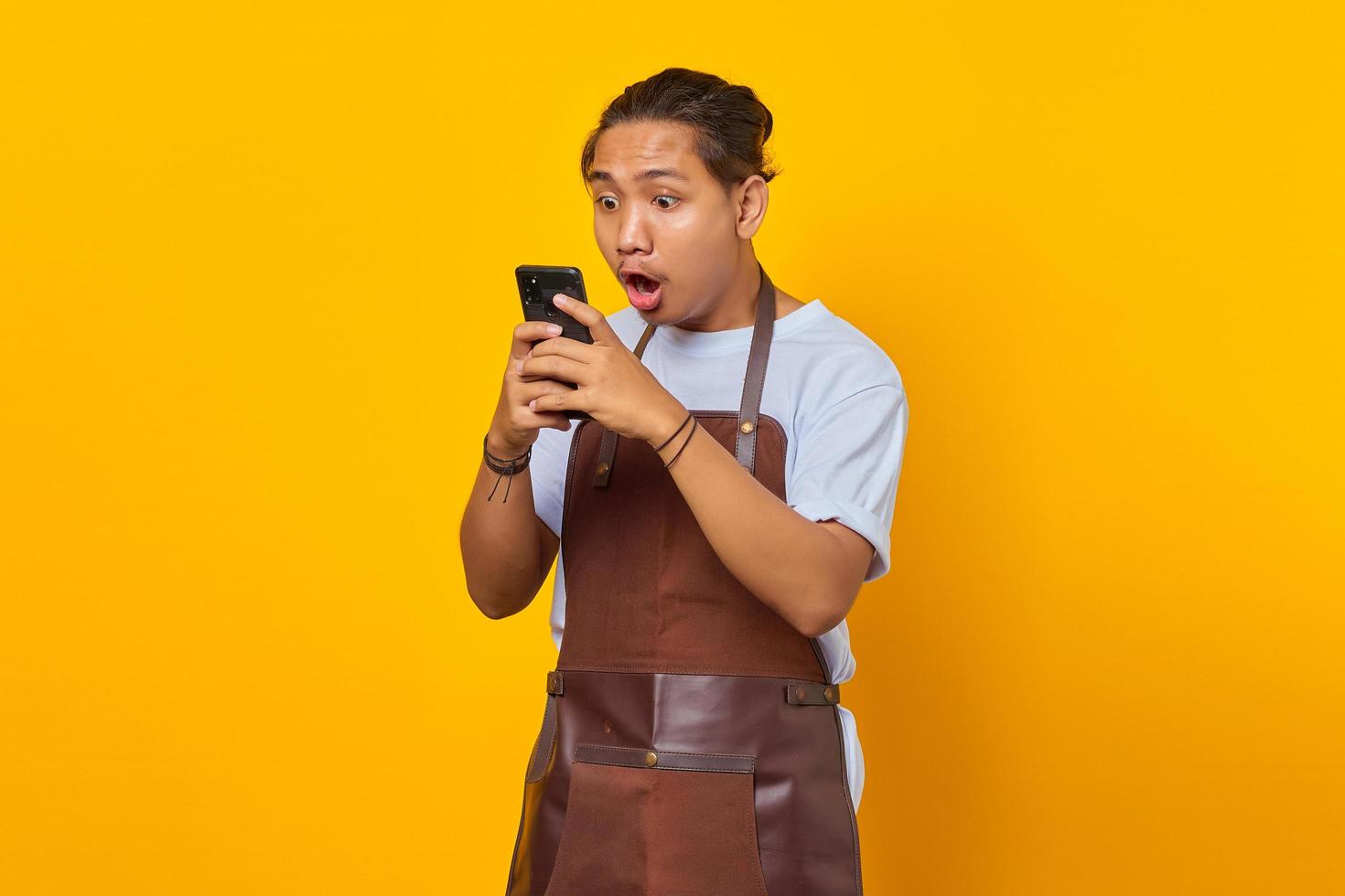 Retrato de guapo barista que está conmocionado y sorprendido al ver la promoción de descuento en el teléfono inteligente sobre fondo amarillo foto