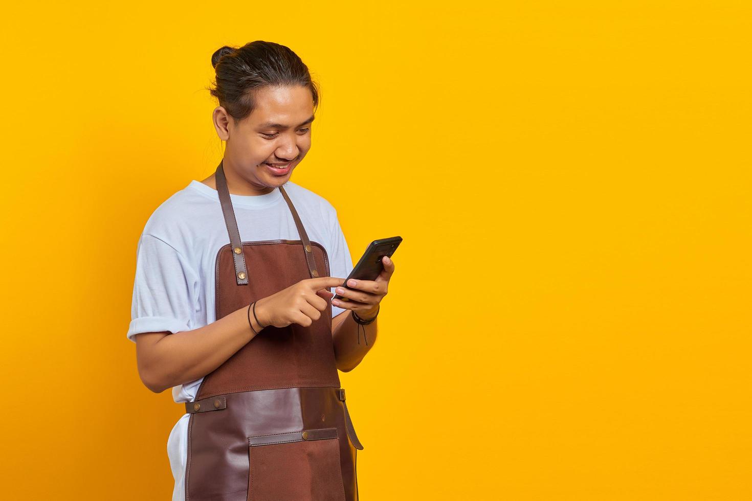 Portrait of Asian young man wearing apron smiling broadly looking at incoming message on smartphone on yellow background photo