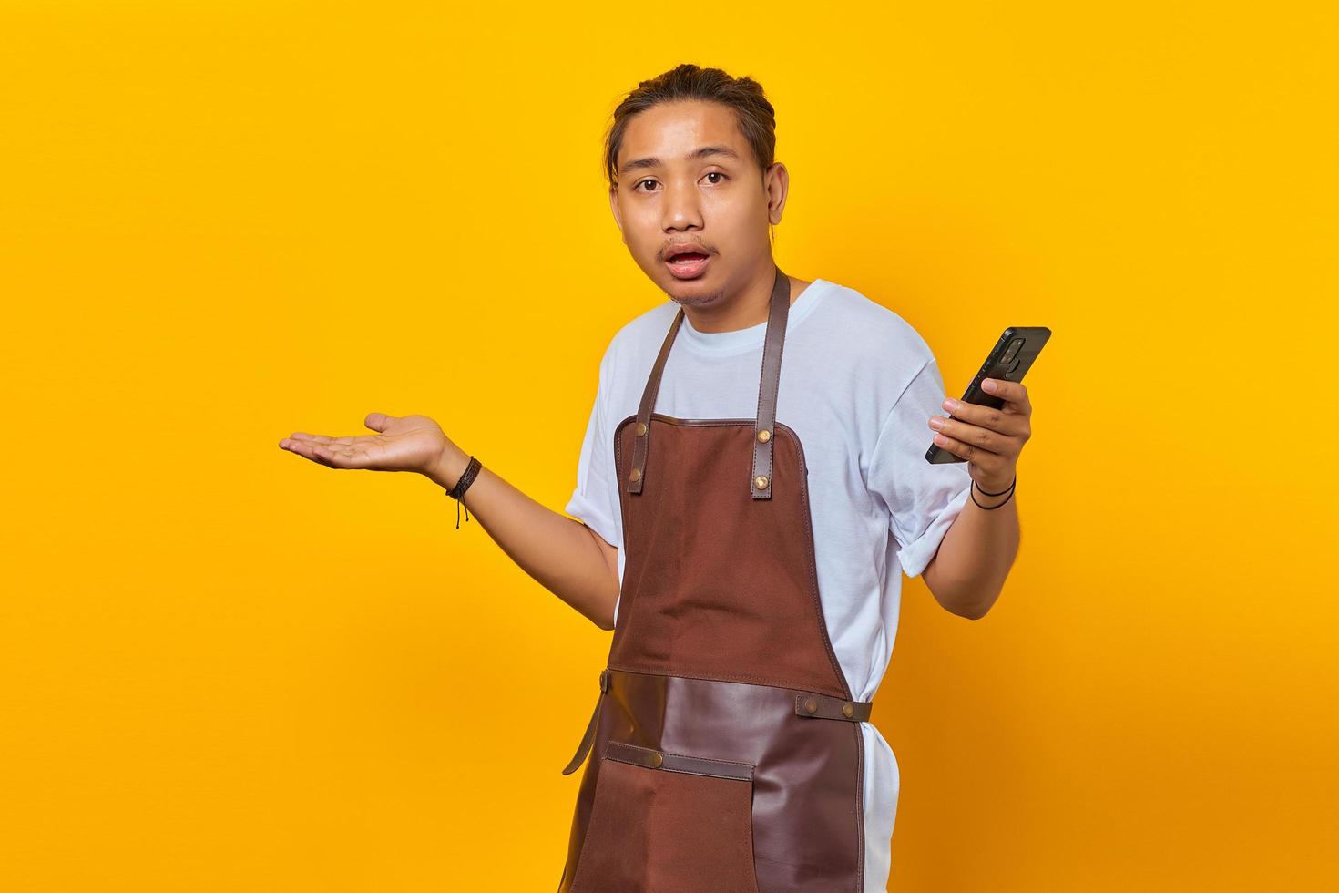 Portrait of surprised handsome Asian young man wearing apron holding smartphone with confused and unhappy expression on yellow background photo