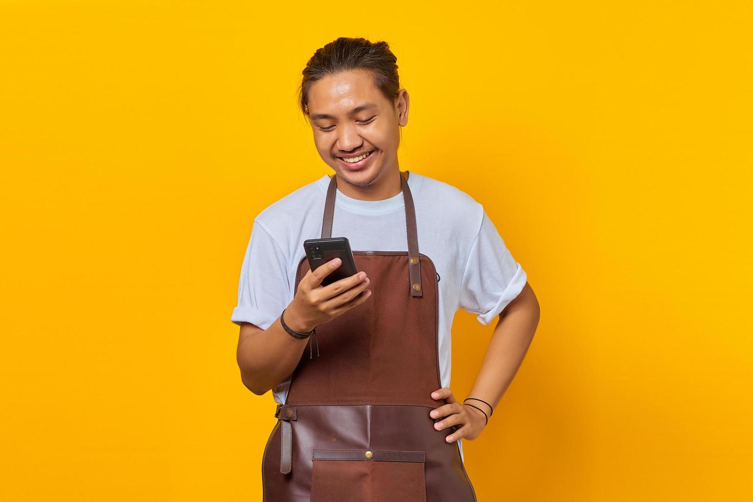 Retrato de un joven asiático vistiendo delantal sonriendo ampliamente mirando el mensaje entrante en el teléfono inteligente sobre fondo amarillo foto