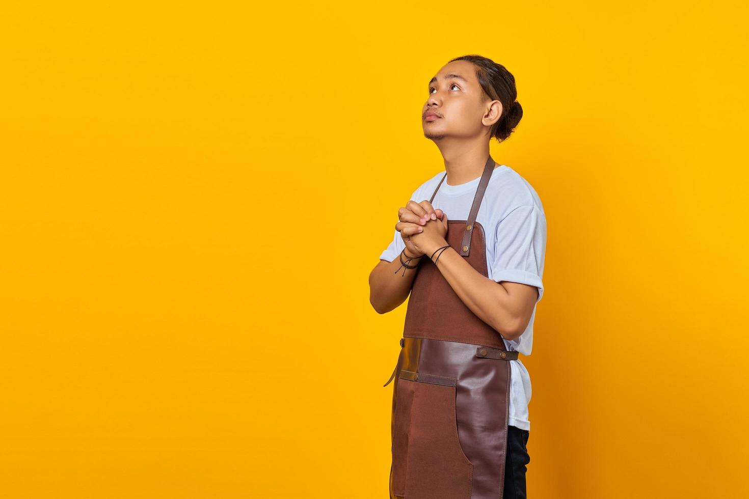 Portrait of handsome Asian young man wearing apron praying, believing in master on yellow background photo
