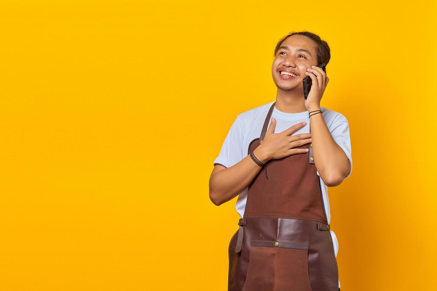 Portrait of cheerful handsome Asian young man wearing apron talking on mobile and hands on chest isolated on yellow background photo